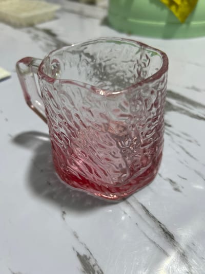 An extreme close-up shot focuses on a small, textured glass mug with a pinkish hue. The mug has a handle and is partially filled with a red liquid. It sits on a white surface with a marble-like pattern. The background is blurred and shows hints of other objects, including what appears to be a light green container. The lighting appears to be natural or from an overhead source, casting a shadow from the mug to the left. There are no people visible in the image, and the scene is static. The specific location in Дедемель, Kyrgyzstan cannot be determined from the visual information provided in the image.