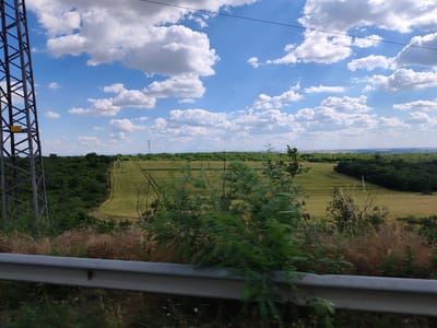 The image depicts a wide, rural landscape under a bright, cloudy sky. In the foreground, a metal guardrail runs horizontally across the bottom of the frame, partially obscured by dry grasses and green foliage. To the left, a large, dark metal lattice tower stands prominently, its structure detailed with crisscrossing beams. A yellow warning sign is affixed to the tower.
Beyond the immediate foreground, the scene opens up to a vast, green field with visible tire tracks suggesting agricultural activity. This field is bordered by dense, dark green trees that form a natural boundary. In the distance, the landscape rolls gently, with more trees and fields stretching towards the horizon. The sky is a vibrant blue, dotted with fluffy white cumulus clouds, indicating fair weather. The lighting suggests it is daytime, likely late morning or early afternoon.
The overall impression is one of peaceful, open countryside. There are no people visible in the image. The setting appears to be a rural area in Zgalevo, Bulgaria, as indicated by the context. The image captures a moment of natural beauty and agricultural expanse, likely taken from a moving vehicle due to the slight blur in the foreground vegetation.