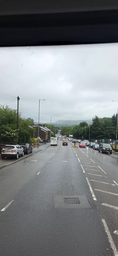The image shows a street scene on an overcast day, likely in Tameside, United Kingdom, judging by the architecture and road markings. The road is wet, indicating recent rain. Cars are parked on the left side of the road, and traffic is moving in both directions, though it appears congested. Buildings line the left side of the road, with trees and greenery beyond. In the distance, hills are visible under a cloudy sky. Several streetlights are present along the road. The overall impression is of a suburban or semi-rural road on a cloudy day.