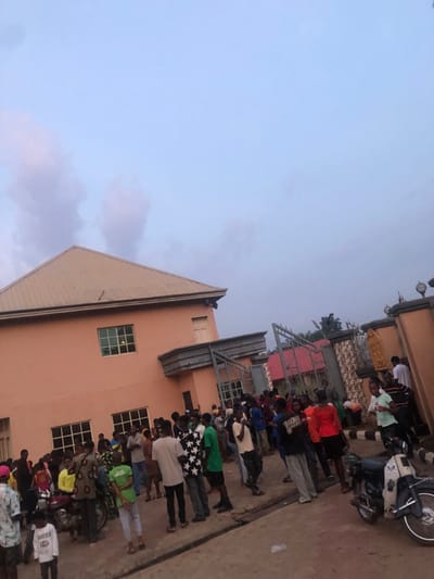 This image depicts a crowded scene on a paved outdoor area in Isiamigbo, Nigeria, during what appears to be late afternoon or early evening. The dominant structure is a two-story building with a peach-colored facade and a light brown hip roof, featuring multiple rectangular windows. To the right of this building is an open metal gate set within a patterned block fence, which includes a decorative golden relief or statue on one of its pillars.
Approximately 40-50 individuals, primarily young adults, are gathered across the foreground and midground. They are dressed in casual attire, including t-shirts, shirts, and trousers of various colors and patterns. Most individuals are standing, appearing to mill about or observe their surroundings. A few are engaged in conversation. A light-colored motorcycle is parked in the lower right corner, facing left, and a yellow motorcycle is partially visible among the crowd in the lower-left midground. The sky above is a pale blue with scattered, light-colored clouds. Beyond the gate and fence, distant trees and other structures are partially visible.