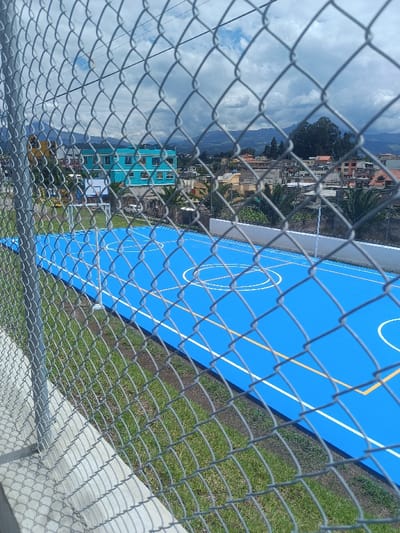 The image displays an outdoor, fenced-in sports facility located in Sangolquí, Ecuador. The foreground is dominated by a grey chain-link fence, through which the scene is viewed, with a concrete barrier and green grass visible at its base. Directly beyond the fence, a vibrantly blue multi-sport court is visible, featuring white and yellow painted lines delineating areas for basketball (including a key and center circle) and potentially other sports like volleyball or tennis. A white basketball hoop with a backboard stands at the far end of the court, and a small white soccer goal is positioned near the left edge. No individuals are present on the court or within the immediate facility area.
In the mid-ground and background, an urban residential area unfolds under a cloudy sky. Prominently featured is a multi-story turquoise building with white-framed windows, situated behind the sports court. Surrounding this are numerous other residential structures of varying colors and architectural styles, interspersed with palm trees and general vegetation. Distant mountains are visible on the horizon, partially obscured by the clouds. The overall environment suggests an institutional or public recreational area within a populated city.