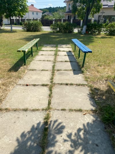 A daytime, eye-level, outdoor shot shows a paved walkway with grass growing through the cracks, leading towards two park benches. The bench on the left is painted green and the bench on the right is painted blue. Both benches are simple, slatted wooden designs with metal legs. They are positioned on either side of the walkway, facing outward. In the background, there are trees, houses, and a fence. The scene appears to be in a residential area or park in Ruseștii Noi, Moldova. The sky is blue with some scattered clouds. Shadows from trees are visible on the walkway and grass.