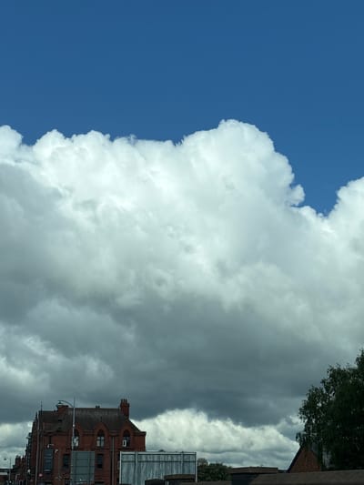 The image shows a large, white cumulus cloud against a blue sky. A red brick building is visible in the lower right corner, appearing to be The Crewe Arms Hotel in Crewe, United Kingdom. Some trees are seen above the building. The image orientation is vertical.