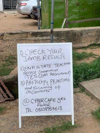 The image shows a white board sign with black marker handwriting, positioned on a dirt path, at the location of Latitude: 7.252, Longitude: 5.199 in Ondo, Nigeria. The sign advertises the services of a cyber cafe, located on the 3rd floor, with a phone number listed for contact. The sign lists three services: 1) Checking JAMB results, 2) Ekiti State Teaching Service (Commission) TES COM Recruitment, 3) Photocopy, printing, and scanning of documents. The sign is leaning against a low wall, partially obscured by green grass. The background shows a green painted metal shed and a sliver car parked on a dirt driveway. The sign appears to be aimed at pedestrians walking along the path.
