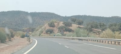 The image shows a highway curving through a hilly, arid landscape with scattered trees and brush. A guardrail runs along the right side of the road. Two "80" speed limit signs are visible on posts on the left side of the road, one further in the distance than the other. A dark-colored car is parked near the speed limit signs. The sky is overcast and light. The terrain suggests a location in a dry, Mediterranean climate.