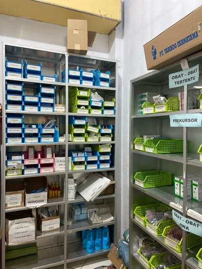 The image depicts a meticulously organized storage area, likely a pharmacy or medical supply room, in Margoyoso, Indonesia. Shelves are filled with various medical supplies, neatly categorized and labeled. Prominently displayed are boxes and containers of medications, including bottles labeled "OBAT - OBAT TERTENTU" (Certain Medicines) and "OBAT - OBAT JIWA" (Psychiatric Medicines). There are also trays filled with items such as syringes, medical tubing, and other disposable medical equipment.
Green and blue plastic baskets are used to hold smaller items, and many containers are labeled in what appears to be Indonesian, with terms like "PREKURSOR" (Precursor), "TRAMADOL", "OCTENIC SPRAY", and "GLOVECLEAN". Larger cardboard boxes, some with company logos such as "PT. YARINDO FARMATAMA", are also visible. The lighting suggests an indoor setting, possibly during daylight hours given the overall brightness. The scene conveys a sense of order and preparedness, common in a healthcare or pharmaceutical environment. No people are visible in the image.