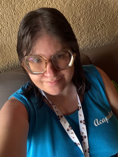 A person, likely female, is captured in a close-up selfie, reclining indoors. They have dark brown hair, which appears slightly damp. Large, clear-framed or light brown-framed glasses are worn, reflecting light from an unseen source. The person looks directly at the camera with a neutral to slightly pleasant expression, revealing their brown eyes, nose, and part of their mouth with light pink lips.
They are wearing a light blue t-shirt with the word "Acapulco" embroidered in white script on the left chest area. The t-shirt fabric shows some pilling or texture. Around their neck is a white lanyard patterned with small, cartoon-style images of Harry Potter, sorting hats, and golden snitches.
The background consists of a textured, golden-brown surface, possibly a stucco wall or heavily textured wallpaper, behind their head. A dark, possibly leather or fabric, armrest or cushion is visible behind their back and shoulder. The lighting suggests an indoor setting. The "Acapulco" shirt and the location context "Tres de Mayo, Mexico" suggest a connection to Mexico.