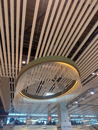 The image depicts the interior of a modern commercial space, likely a shopping mall or airport terminal, featuring a prominent, large circular chandelier with numerous clear, dangling elements. The chandelier is suspended from a ceiling composed of parallel white slats, creating a textured, angled effect. The space below the chandelier shows people walking on a staircase and congregating in a seating area, with signage visible, including the word "ELECTRONICS" on a white pillar. The overall environment is well-lit and suggests a public, indoor setting. The location is identified as Bubuieci, Moldova.