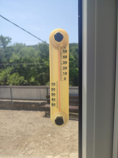 A vertical yellow thermometer is mounted on a window. The thermometer displays a temperature of approximately 32 degrees Celsius. The background shows a clear blue sky, green trees, and a fence. The image is taken from inside a building looking out.