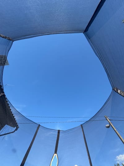 The media file shows a view looking upwards through a dark blue safety net enclosure against a clear blue sky. The net forms a roughly square opening in the center, with the edges of the net visible in the foreground. The net appears to be made of a mesh material, allowing the sky to be seen through it.
In the background, elements of a building with a brick facade and a corrugated roof are partially visible beneath the net. A thin, metallic lightning rod or antenna is also visible on top of the roof. The lighting suggests it is daytime, likely under sunny weather conditions. The overall impression is of being inside or under a structure with protective netting, looking out at the open sky. There are no people visible in the media file. The setting suggests an outdoor area, possibly a sports facility or construction site where safety netting is used. The lack of prominent shadows might indicate a time close to midday. No visible text is present.
