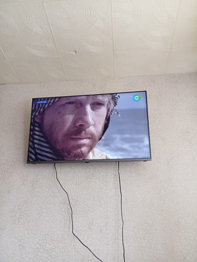 A television screen mounted on a textured, light-colored wall displays a close-up of a man's face. The man has fair skin, a beard, and is wearing a dark and white patterned scarf around his head. A blue logo with a green circle and the word "POWER" is visible in the upper right corner of the screen. A black cable runs from the bottom of the television down the wall. The ceiling above the wall is textured with repeating circular patterns.