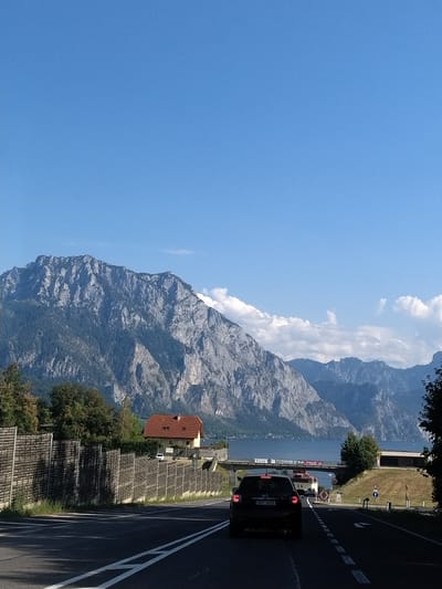 The image captures a scenic view of the Traunsee lake and the surrounding mountains, specifically the Traunstein, as seen from the road leading into Gmunden, Austria. In the foreground, a black car with license plate 5K5 3233 is driving on a paved road with white lane markings. A wooden fence runs along the right side of the road. A red-roofed house with light yellow walls is visible behind the fence. Further in the distance, the Traunsee lake is visible, with boats dotting its surface. The imposing Traunstein mountain dominates the background, its rocky slopes covered in dark green trees. The sky is a clear blue with a few scattered white clouds. The overall impression is a picturesque landscape scene, likely captured on a sunny day.