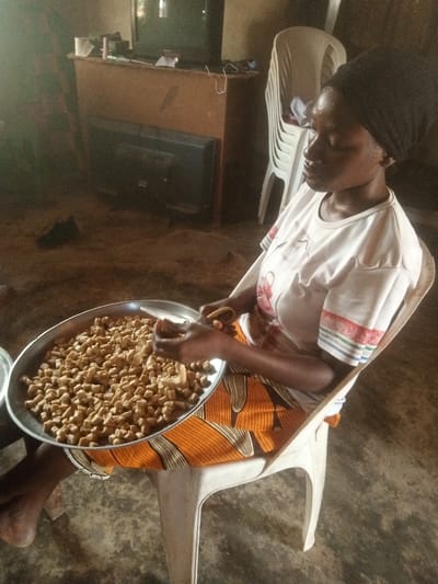 In Gudige Sabo, Nigeria, an indoor scene depicts a woman engaged in food preparation. The subject, a woman with dark skin, wears a black headwrap, a white short-sleeved t-shirt with striped cuff details, and an orange and black patterned wrap skirt. She is seated on a white plastic chair. A large, circular, silver-toned metal tray rests on her lap, filled with numerous irregularly shaped, light brown, uniform-sized snack pieces. Her hands are positioned over the tray, actively handling the snack pieces, with what appears to be a shaping or breaking action involving a small tool or another piece of the food item. Perspiration is visible on her forehead and face. The background includes a wooden media console supporting an older model television on top and a smaller flat-screen television below. Wires and miscellaneous items are present on the console. A stack of white plastic chairs stands to the right. The floor is a textured, unpaved earth surface, and the walls are plain and unadorned. Ambient lighting illuminates the scene from an unseen source to the left.