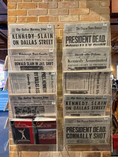 A two-tiered display of newspapers commemorating John F. Kennedy's assassination. Titles include "The Dallas Times Herald" with the headline "President Dead, Connally Shot," "New York Herald Tribune," and "Fort Worth Press," highlighting the national impact. "The Dallas Morning News" appears several times, detailing Kennedy's slaying and Johnson's succession. One newspaper reports Oswald's death in jail. A stack of books titled "Legacies" is seen on the bottom right, and everything is neatly encased in clear plastic holders, mounted on a brick wall.