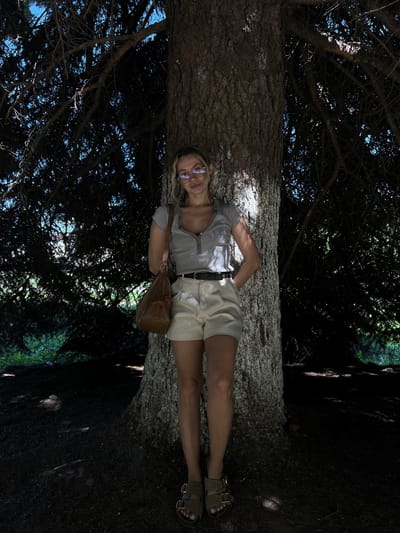A woman with blond hair and glasses lies on a large fallen tree trunk in an outdoor setting. She wears a gray shirt, khaki shorts, sandals, and a belt, and carries a brown purse. The tree trunk occupies the central space with a dark green and black forest environment surrounding the scene. The location is implied to be in the city of Слобода, Russia.