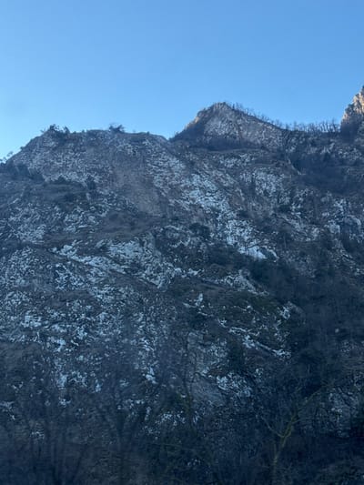 A steep, rocky cliff face, mottled with patches of white, likely snow or ice, rises dramatically against a clear blue sky. Sparse trees and vegetation cling to the cliff's edge. The rock formation is complex, with varying textures and shadows defining its structure. The overall perspective is looking upward at a significant elevation.