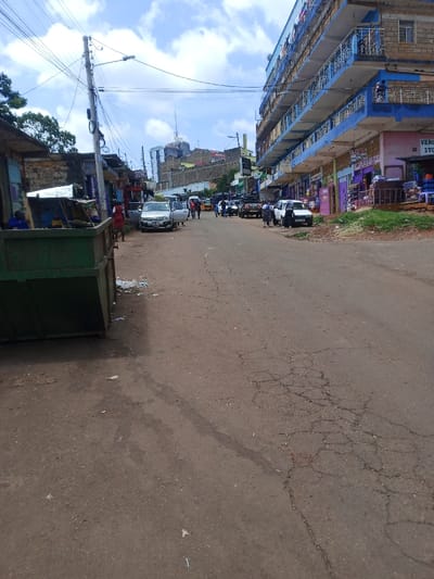 The image displays an urban street scene in Murang'a, Kenya, under a partly cloudy sky. The foreground features an unpaved road with visible cracks, dirt patches, and minor debris. A large green metal skip/dumpster is positioned on the left.

On the left side of the road, several makeshift stalls constructed from stone and corrugated iron are visible. Two individuals, one in a blue shirt, are seated near these stalls. A person in a red top stands near a light-colored sedan with an open driver's door, parked on the left shoulder.

The midground shows multiple vehicles, including sedans and a yellow auto-rickshaw, parked or moving along the road. Numerous pedestrians are dispersed across the street and sidewalks.

The right side of the scene is dominated by a multi-story building featuring stone and blue painted sections with blue balcony railings. The ground floor houses retail businesses with visible signage, including a partially legible "VERONA STORE."

In the background, an elevated section of the city is visible, populated with additional buildings and topped by a communications tower. Overhead utility lines are prominent, supported by a utility pole in the mid-left.