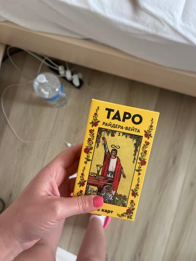 An overhead photograph, taken in Heusden-Zolder, Belgium, depicts a human hand holding a yellow tarot card box. The hand, exhibiting pink nail polish on the visible thumb, grasps the box firmly. The box's front displays the Russian text "ТАРО РАЙДЕРА-ВЕЙТА" (Tarot Rider-Waite) in black capital letters above an illustration of The Magician tarot card from the Rider-Waite deck. The card image depicts a robed figure with an infinity symbol above its head, holding a wand and standing behind a table with a cup, pentacle, and sword. The illustration is framed by a decorative border of red roses and green foliage. Below the illustration, the partial Russian text "3 карт" is visible. The background, out of focus, shows a light-colored wooden bed frame to the left and top. White, rumpled bedding is present on the bed. On the light-colored wooden floor, a clear plastic water bottle with a blue label is visible along with several white tangled cables. A black power adapter with white plugs is partially obscured beneath the bed frame.