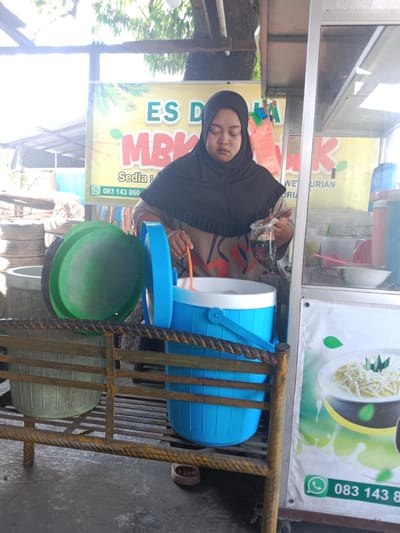 In Sarimulyo, Indonesia, a woman appearing to be in her 20s or 30s operates an outdoor street food stall. She wears a black hijab and a light-colored, patterned long-sleeved top, with her eyes closed or looking downwards, focusing on her task. Her left hand holds a clear plastic bag containing a dark, layered beverage or dessert. Her right hand, holding an orange ladle, is extended into a large, open, blue insulated container, from which she is scooping a white substance. This container rests on a low, rusty, yellow-painted metal rack. To the left of the blue container, a large green plastic basin with its lid ajar is also positioned on the metal rack.
The stall features a prominent yellow banner with red text reading "ES DURIAN MBK" (Es Durian Mbak), beneath which is "Sedia : WE URIAN". A green WhatsApp icon and phone number "083 143 860" are printed on this banner. To the woman's right, a glass or clear plastic display case shows various clear plastic bowls and containers within. A white poster is affixed to the lower part of this display case, featuring an image of a dessert with a white shredded topping and green leaves, along with a partial WhatsApp icon and number "083 143 8". In the background, to the far left, a stack of worn-out tires is visible. The scene is brightly lit by daylight, casting some overexposure in the upper left. The woman's light-colored sandals are visible on the concrete ground beneath the stall.