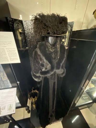 The image shows a museum exhibit. A mannequin is dressed in a black Victorian-era gown with a fur-trimmed coat and a large feathered hat. The exhibit is in a dimly lit room with a black background. There is a display case with artifacts on the right side of the image. On the left side of the image, there is a description of the exhibit, which appears to be related to the life and etiquette of St. Petersburg in the late 19th century. There is no indication that this exhibit is located in the city of Voskhod, Russia.