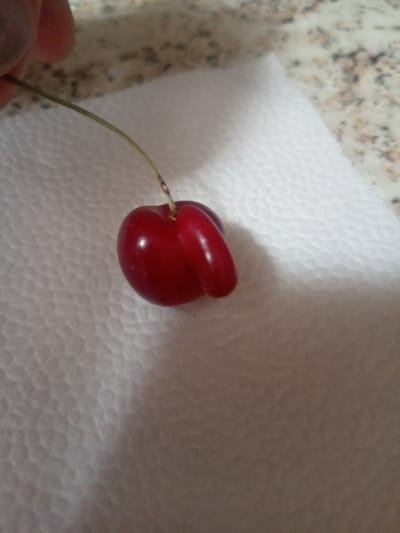 A single, slightly deformed cherry with a stem rests on a white paper napkin. The cherry is dark red and appears shiny, with a prominent protrusion on one side, giving it an unusual shape. The napkin has a textured surface and is placed on a speckled countertop, likely granite or marble, which exhibits shades of brown, gray, and white. A person's hand, with skin tone variations, is partially visible in the upper left corner, holding the stem of the cherry. The lighting suggests an indoor environment with a light source casting subtle shadows. There is no indication of the city of Labinsk, Russia, or any specific urban context in this image.