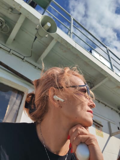 A woman with light brown hair is looking to the right, wearing glasses and an earbud. She has her hand resting on her chin. She is on a boat with a blue sky and white clouds in the background. The boat appears to be docked or moving slowly. The scene is well-lit by natural sunlight.