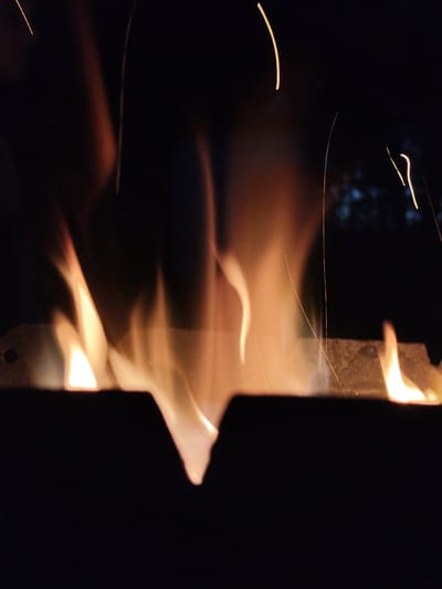 In the city of Kolpakovo, Russia, a close-up, low-angle shot captures a roaring fire at night. The flames, in shades of orange and yellow, dance against a dark background, with thin sparks ascending into the darkness. A metal grate or trough, possibly part of a brazier or fire pit, is visible at the bottom of the frame, with flames emerging from its edges. The overall impression is one of intense heat and motion within a confined space.