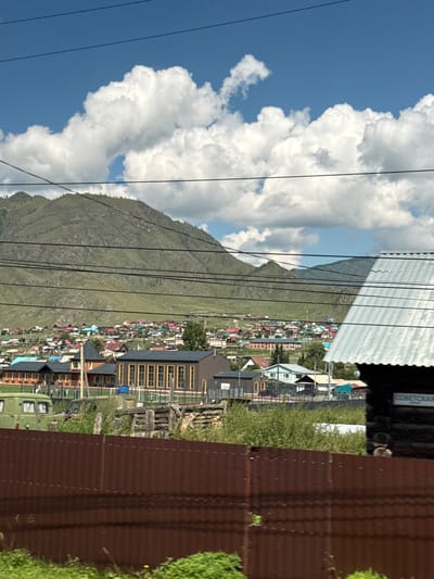 In Onguday, Russia, a village lies nestled at the foot of a lush green mountain under a bright blue sky dotted with fluffy white clouds. The village is comprised of numerous houses with colorful roofs, creating a vibrant patchwork. In the foreground, a rusty corrugated metal fence runs horizontally, partially obscuring the view of the village. Behind the fence, there are some wooden structures and a green vehicle. Further back, a dark wooden building with a corrugated metal roof is visible on the right side of the frame. The overall scene depicts a rural residential area in a mountainous region.