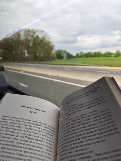 A person is reading a book while apparently traveling on a highway, as evidenced by the passing scenery through the car window. The book is open to two pages with text in Bulgarian, titled "Петдесет и пета глава Том" ("Fifty-fifth Chapter Tom").
Outside the car window, the landscape consists of trees and green fields under an overcast sky. The highway appears to have multiple lanes. Based on the style of the road infrastructure and signage, the location is likely the M1 motorway near Milton Keynes in the United Kingdom.