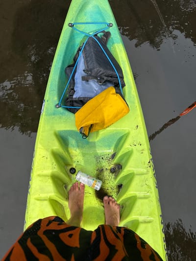 An overhead shot captures a pair of bare feet inside a bright lime green and yellow kayak, floating on dark, calm water. The kayak appears to be a sit-on-top model, with the brand name "OCEAN KAYAK" partially visible along the side. The interior of the kayak is marked with some dark dirt and debris, particularly around the foot area.
Resting in the front of the kayak is a black and blue backpack, secured by a bright blue bungee cord system. Next to the backpack is a yellow dry bag. Near the person's feet, a white beverage can with colorful graphics is visible, displaying the text "CELSIUS", "ESSENTIAL ENERGY", and "ZERO SUGAR". The can also indicates "GREEN TEA &..." and other small, illegible print.
The person's feet are positioned towards the rear of the kayak, and the bottom of their orange and black tiger-print swim shorts is visible on the right side of the frame. A portion of an orange kayak paddle blade is seen in the water, just beyond the front of the kayak. The dark water reflects distorted images of trees and sky, suggesting an outdoor setting like a river or calm lake. The scene depicts a moment of rest during a kayaking activity in Tampa, United States.