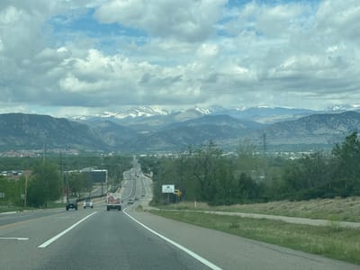 The image presents a view from a road heading towards the city of Boulder, Colorado, United States. The foreground is dominated by a two-lane road with a dividing line. Three vehicles are visible on the road: a dark-colored pickup truck, a white SUV, and a box truck displaying an advertisement for "Whole Mountain Meals." A sign indicating "Traffic Laws Photo Enforced" stands on the right side of the road. Beyond the road, the landscape transitions to grassy areas and scattered trees. In the middle ground, the city of Boulder is visible with buildings and a dense tree canopy. The background features a mountain range with snow-capped peaks. The sky is overcast with scattered clouds.