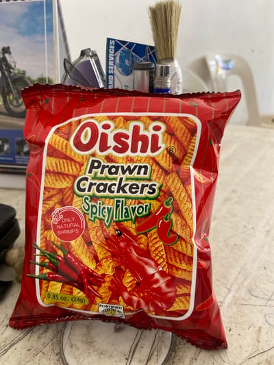 The image shows a bag of Oishi Prawn Crackers, Spicy Flavor, placed on a textured surface, possibly a table or countertop. The bag is predominantly red with yellow and green accents, featuring illustrations of crackers, chili peppers, and a shrimp. Text on the bag indicates the product name, flavor, and weight (0.85 oz. / 24g). The background is blurred but suggests an indoor environment with objects like a motorcycle magazine, a brush, and a sink faucet visible. This scene is located in San Jose del Monte, Philippines, a city known for its residential areas and commercial establishments.