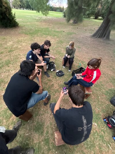 From an elevated angle, a group of seven people are casually gathered on grassy ground. One woman wearing a red jacket is seated on a crate and focused on a phone. Another person in muted clothing is seated on a similar crate. A cluster of three people is closely interacting on the ground. One man is kneeling, using his phone. Another man's legs are visible in the bottom of the frame. A coil of wire rests near the group. Gear is visible in the upper corner.