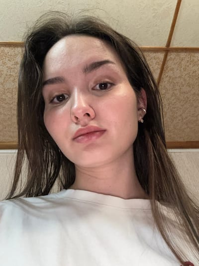 This image displays a single individual, an adult female, located indoors in the city of Bayronovka, Russia. The subject is captured from a slightly low-angle perspective, looking directly at the camera. She has long, dark brown hair falling over her shoulders and is wearing a plain white crew-neck top. Her visible right ear features two pieces of jewelry: a small, silver-toned hoop earring in the upper helix and a small, dangling earring with a clear element in the lobe. Her facial features include dark eyes and well-defined eyebrows. The background comprises two distinct sections: a light-colored wall with a subtle floral or abstract pattern, and a lighter-toned ceiling featuring a grid-like design created by wooden beams.