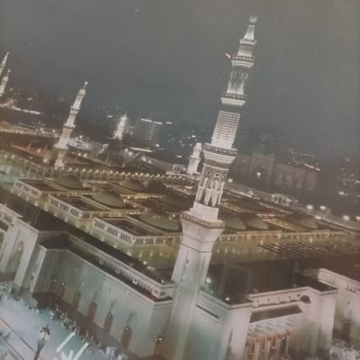 This image presents an aerial night view of a large, brightly illuminated mosque complex situated in the city of Pasir Muncang, Indonesia. The primary subject is a sprawling religious edifice characterized by multiple white or light-colored buildings and distinctive architectural elements. In the immediate foreground on the right, a towering, ornate minaret stands prominently, showcasing intricate tiered designs and illuminated balconies. A second, similarly designed minaret is visible further back on the left side of the complex. The mosque features numerous flat-roofed sections and a central area distinguished by multiple circular or domed roofs. Extensive courtyards and open areas within the complex are clearly demarcated by rows of bright ground-level lights, outlining pathways and architectural perimeters. Small, indistinct figures, indicative of a gathering or movement of people, are visible along the lower-left edge of the complex, suggesting human activity. The background reveals a vast urban landscape extending under a dark night sky, marked by numerous illuminated structures and city lights, indicating a large population center. The environment is dark save for the extensive lighting of the mosque and surrounding city.