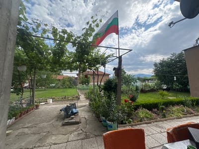 The image depicts an outdoor scene in the city of Zlatovrah, Bulgaria. A section of a patio with an orange chair and tiled flooring is visible in the upper left. In the background, there's a well-maintained garden with various flowers and greenery. A utility pole bisects the image. A flag with green, white, and red stripes (likely the Bulgarian flag) is hanging below a vine-covered structure. The sky is overcast with many white clouds.