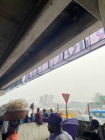 The image captures a street scene under a concrete overpass in Rumuokoro, Nigeria, during overcast weather, with evidence of rain. The upper frame is filled by the grey underside of a concrete overpass structure, featuring multiple beams and exposed conduits. Water droplets are visibly falling from the overpass structure. Below, a bustling street level is present.
In the foreground, on the left, a woman wearing an orange top carries a large, round, plastic-covered basket filled with items, balanced on her head. To her right, partially obscured, a man wearing a purple baseball cap and a light-colored shirt holds a partially open purple umbrella. Further to the right, a child or young person is visible.
In the mid-ground, numerous individuals are present, some seated on a low concrete barrier, including one person in a red plaid shirt. Vehicles, including a yellow commercial bus, are visible on the street, indicating active traffic. In the background, a large billboard displays an image of a man in a suit and text. A prominent red triangular "GIVE WAY" sign is visible, along with a smaller white sign that includes "RUMUOKORO" in its text, confirming the location. The overall lighting is diffused, consistent with an overcast sky.