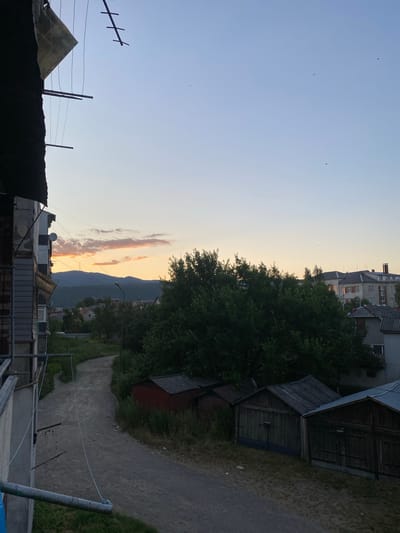 This is a photograph taken in Khust, Ukraine, likely during the late afternoon or early evening, given the soft, warm light illuminating the sky and the clouds. The image is oriented vertically, presenting a view of a somewhat rural or semi-urban environment.
In the foreground, on the left, are the dark silhouettes of rooftops and what appear to be structures, possibly outbuildings or sheds. An antenna is visible against the sky. Moving towards the center and right of the image, we see a dense cluster of green trees. Behind the trees, a multi-story building with a grey facade and windows is visible, suggesting residential or communal buildings. Further to the right are several weathered wooden sheds or garages with corrugated metal roofs, situated along a gravel path.
The background is dominated by a mountain range, its slopes covered in dark greenery. The sky transitions from a pale blue at the top to a soft, creamy yellow-orange near the horizon, with streaks of pink and orange clouds indicating the setting sun. A few birds are visible as small specks in the sky. The overall atmosphere is serene and peaceful, capturing a quiet moment in a provincial setting. There is no visible text in the image.