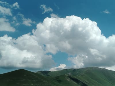 The image shows a scenic landscape of green mountains under a bright blue sky filled with dramatic white clouds. The mountains are covered in lush green vegetation, with some rocky outcrops visible on their peaks. The terrain appears to be rolling and undulating, suggesting a mountainous region. The sky is a vibrant blue, with a large, fluffy cumulus cloud dominating the upper center of the frame. Other smaller clouds are scattered across the sky. The lighting suggests it is daytime, likely late morning or early afternoon, with the sun casting shadows on the mountains, highlighting their contours. No people or man-made structures are clearly visible in the image, emphasizing the natural beauty of the scene. The overall impression is one of vastness and natural grandeur. This scene is characteristic of the natural beauty found in the region of Tsilkar, Armenia, known for its mountainous terrain and picturesque landscapes.