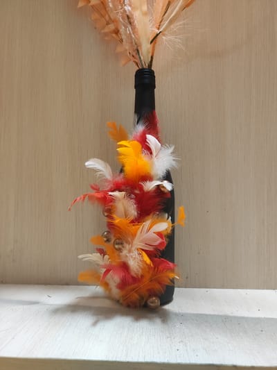 A black bottle decorated with an assortment of colorful feathers in shades of red, orange, and yellow, as well as small golden beads, stands on a white wooden surface against a textured beige wall. The bottle is topped with dried, light-colored decorative plumes. The image is captured in a close-up, still-life composition. The location is not specified as Алабино, Russia, as this information is not present in the image or its context.