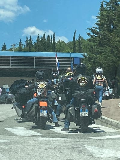 A group of motorcyclists are parked on a paved surface, possibly a parking lot, in front of a building with a blue roof and white horizontal lines. The building is bordered by trees. The motorcyclists are wearing leather vests with patches, helmets, and jeans. Some of the motorcycles have patches and stickers on them, and one has a license plate that reads "FL 29329". Another license plate reads "53345". A flag with red, white, and blue stripes is visible in the background. The sky is blue with some clouds.