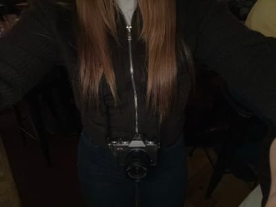 An upside-down view shows a person wearing jeans and a zippered jacket. A vintage camera rests on their torso. Long, brown hair hangs down, partially obscuring the jacket. The person is sitting, surrounded by dark clothing and furniture, creating a slightly cluttered, intimate setting. The image appears to have been taken from above.