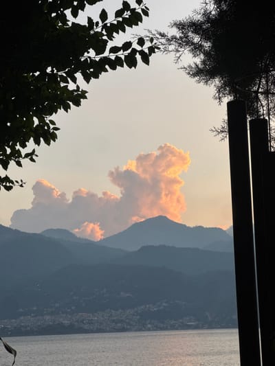 The image captures a serene landscape with distinct layers. Mountains fade into the background, meeting a body of water. A striking, amber-colored cloud formation dominates the sky, its unique shape catching the eye. Tree silhouettes in the foreground add depth, while a shadowed structure peeks in from the top of the frame. The overall composition evokes a sense of peace and natural beauty.