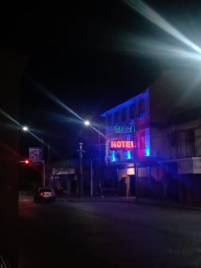 The image depicts a vibrant urban street scene at night in San Fernando, Argentina. A two-story building, presumably a hotel, dominates the right side of the frame, brightly illuminated by striking neon signs. The top sign, in a green or teal color, spells "DELTA," while a larger red neon sign below it clearly reads "HOTEL." The building's facade is also accented with intense blue neon lights along its edges and windows, creating a lively glow.

In the foreground, a dark asphalt street curves slightly. A light-colored car is visible on the left side of the road, heading away from the viewer, with its taillights subtly illuminated. Further down the street, on the left, a red traffic light is active, and several bright streetlights cast strong beams of light with noticeable lens flares across the dark sky. Utility poles and overhead wires are present, characteristic of an urban environment. A smaller, less distinct sign also bearing "HOTEL" can be seen in the background near the traffic light. The clear night sky and the dramatic artificial lighting give the scene an energetic yet calm atmosphere.