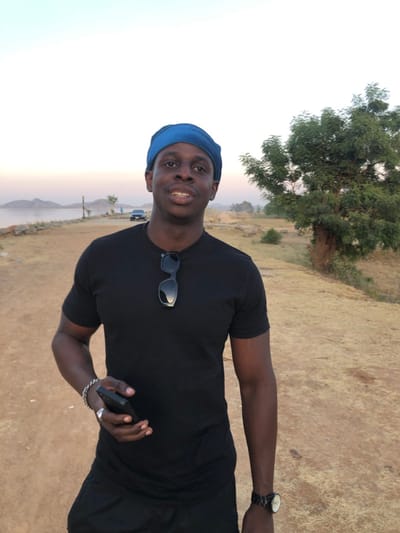 A dark-skinned man wearing a blue head covering and a black t-shirt poses outdoors. He holds a phone in his right hand, with a silver bracelet visible. Sunglasses hang from the front of his shirt. He is wearing a wristwatch on his left wrist. A tree is visible in the background on the left, and a landscape with a distant lake is on the left.