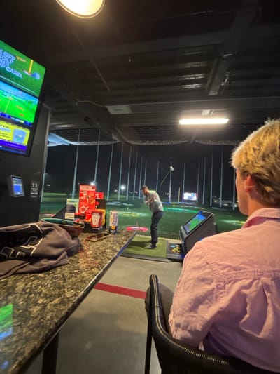 Inside a Topgolf facility, the view is captured from a seated vantage point beside a patterned countertop, displaying condiments, a Corona bottle, and apparel. In the background, a golfer is in mid-swing on the turf. Beyond him, the Topgolf range extends into the night, illuminated by numerous lights and netting. The facility's interior features overhead lighting and a screen showing game information. A person with blond hair and a pink shirt is seated near the camera.
