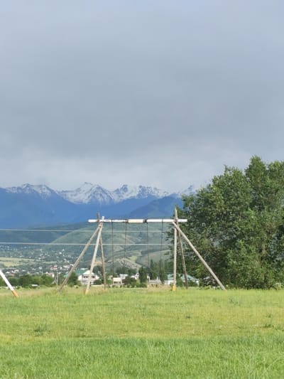 The image depicts a rural landscape with a swing set in the foreground, made of rough wooden poles. In the background, a range of snow-capped mountains forms a dramatic backdrop. Below the mountains, a town or village is visible, with houses nestled amongst green hills. The sky is overcast, with heavy gray clouds dominating the upper portion of the frame. The overall impression is one of a serene, natural setting, with a hint of human habitation in the distance. The location is identified as Dolan, Kazakhstan, a mountainous region known for its natural beauty.
