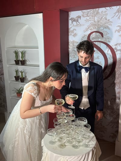 The image captures a wedding celebration at an event venue, possibly in Orizaba, Mexico. A bride in a white lace wedding gown and a groom in a navy suit and bow tie are pouring champagne into a tiered arrangement of champagne flutes on a white-clothed table. The background features a red accent wall and wallpaper with a nature-themed print. A white shelf unit with several small potted plants is also present. The couple appears to be in the act of pouring the champagne, indicating a celebratory toast or tradition.