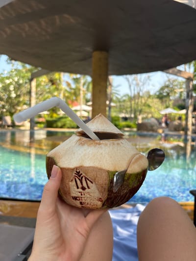 A person holds a fresh coconut with a straw in a light-skinned hand. The coconut has a spoon stuck in the top and a dark brown logo that appears to read "HOTEL BEACH" on the front. The backdrop consists of a swimming pool with a mosaic tile design and green trees and foliage in a tropical setting. The image was taken at The Hot Spring Beach Resort & Spa in Khok Kloi, Thailand.