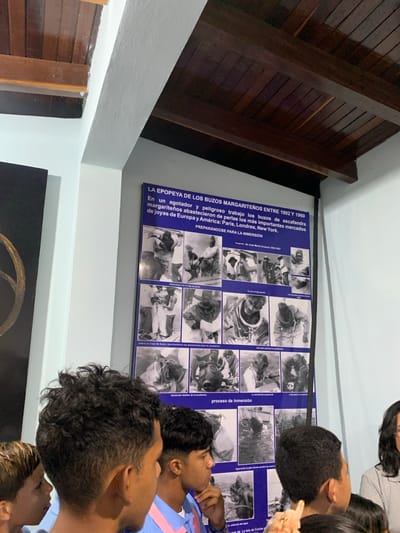 In Boca del Río, Venezuela, a group of individuals, predominantly young males, stand observing a large informational poster mounted on a light blue wall. The poster, titled "LA EPOPEYA DE LOS BUZOS MARGARITEÑOS ENTRE 1902 Y 1960," details the history of divers and their work in pearl fishing. The poster features a grid of black and white photographs illustrating various aspects of diving, from preparation and equipment to the actual immersion process. The ceiling above is constructed of dark wood planks and beams. The foreground is occupied by the heads and shoulders of the observers, some of whom are looking directly at the poster, while others are facing away or looking downwards. The lighting suggests an indoor environment, possibly a museum or cultural center.